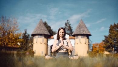 Eine Frau sitzt in der Wiese und macht eine Yoga-Pose. Im Hintergrund sind zwei Türme zu sehen. © Yogafestival Mühlviertel