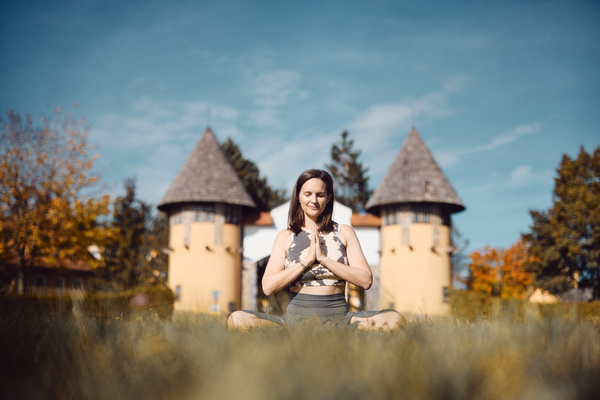 Eine Frau sitzt in der Wiese und macht eine Yoga-Pose. Im Hintergrund sind zwei Türme zu sehen. © Yogafestival Mühlviertel