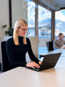 Eine blonde Frau mit Brille sitzt vor einem Laptop und arbeitet.