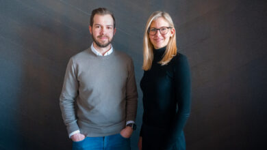 Ein Mann im grauen Pulli und Jeans steht neben einer blonden Frau mit Brille. Beide lächeln.