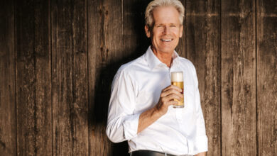 Ein Mann mit einem Bier in der Hand steht vor einer Holzwand und lächelt.