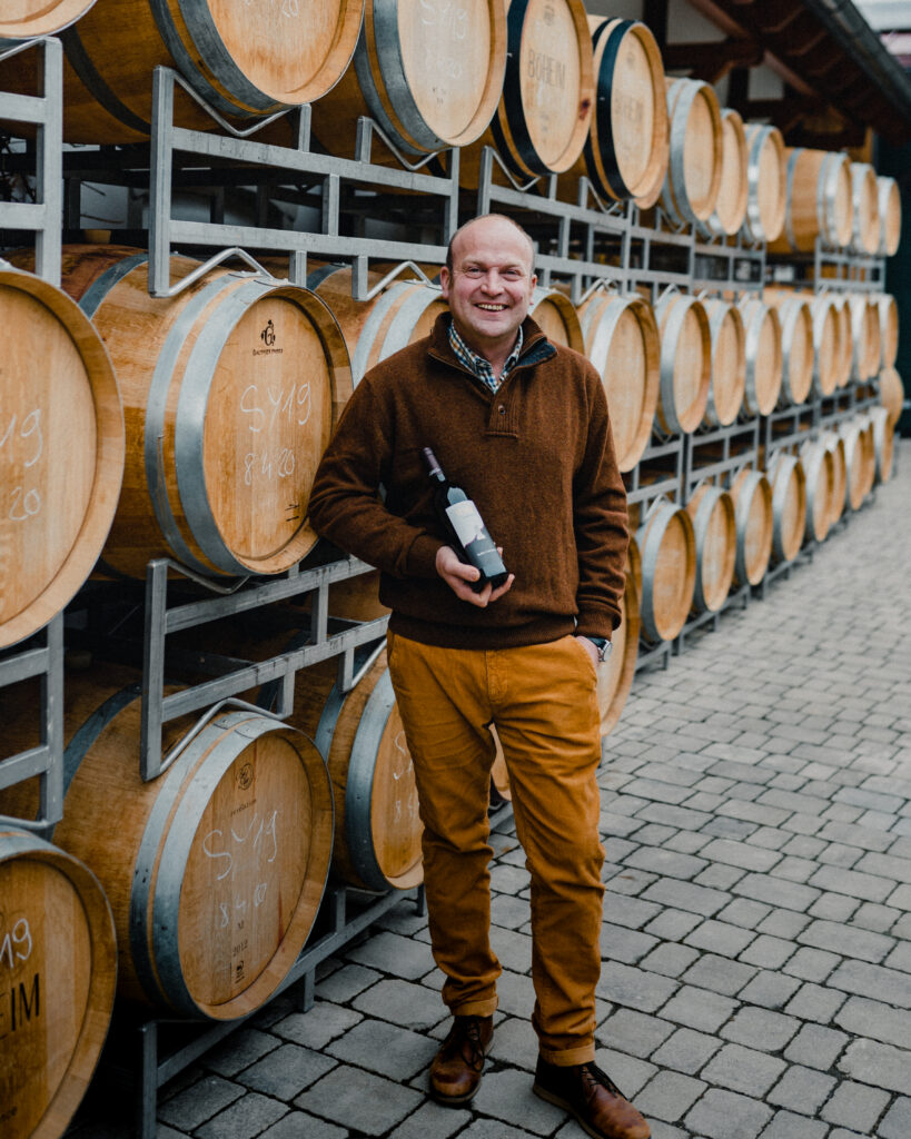 Mann steht vor Weinfässern und hält eine Flasche Wein in der Hand.