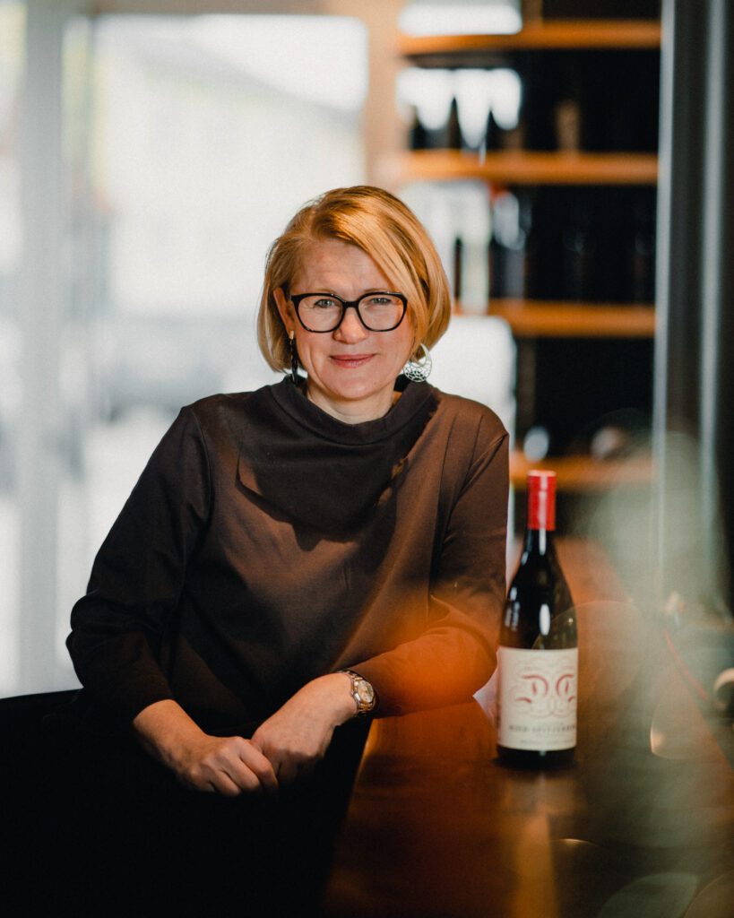 Eine Frau mit Brille und kurzen blonden Haaren steht an einer Bar. Neben ihr eine Flasche Rotwein.