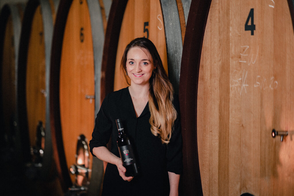 Frau steht vor Weinfässern und haltet eine Flasche Wein.