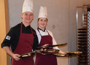 Zwei junge Köche in Uniform halten Teller und lächeln in die Kamera.